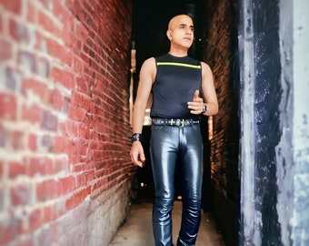 May include: A man wearing a black tank top with a yellow stripe, black leather pants, and black shoes stands in a brick alleyway.