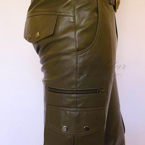May include: Olive green leather cargo pants with a brown leather belt and a zippered pocket on the side. The pants have a flap pocket with two button closures on the back.