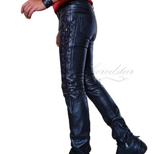 Op de afbeelding: Zwarte leren broek met een veterdetail aan de zijkant. De broek is strak en heeft een rits aan de onderkant van het been. Het model draagt een rood shirt en zwarte schoenen.