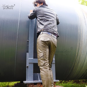 May include: A man wearing a brown leather jacket and olive green leather cargo pants. The pants have multiple pockets and a belt loop. The man is standing next to a large metal cylinder.