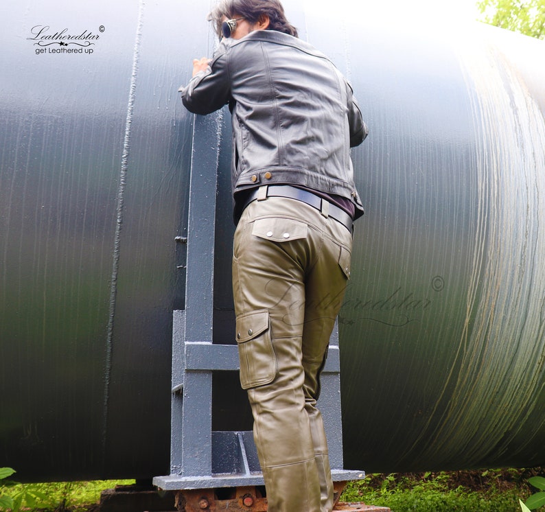 May include: A man wearing a brown leather jacket and olive green leather cargo pants. The pants have multiple pockets and a belt loop. The man is standing next to a large metal cylinder.