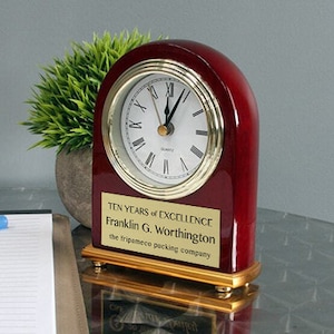 May include: A red wooden clock with a gold trim and a white face. The clock has a plaque on the front that reads "Ten Years of Excellence Franklin G. Worthington the tripumeco packing company".