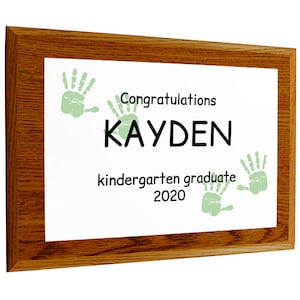 May include: A wooden framed certificate with a white background. The certificate says "Congratulations Kayden kindergarten graduate 2020". There are three green handprints on the certificate.