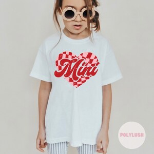 Könnte beinhalten: Weißes T-Shirt mit einem rot-rosa karierten Herz-Design. Das Wort "Mini" ist in einer verblassten roten Schriftart über dem Herzen aufgedruckt. Das Shirt hat kurze Ärmel und scheint aus Baumwolle zu sein.
