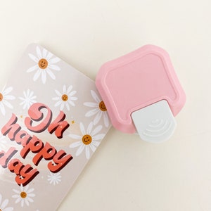 May include: A pink and white paper punch with a daisy design. The punch is on top of a card with the text "Ok Happy Day" in a retro font.