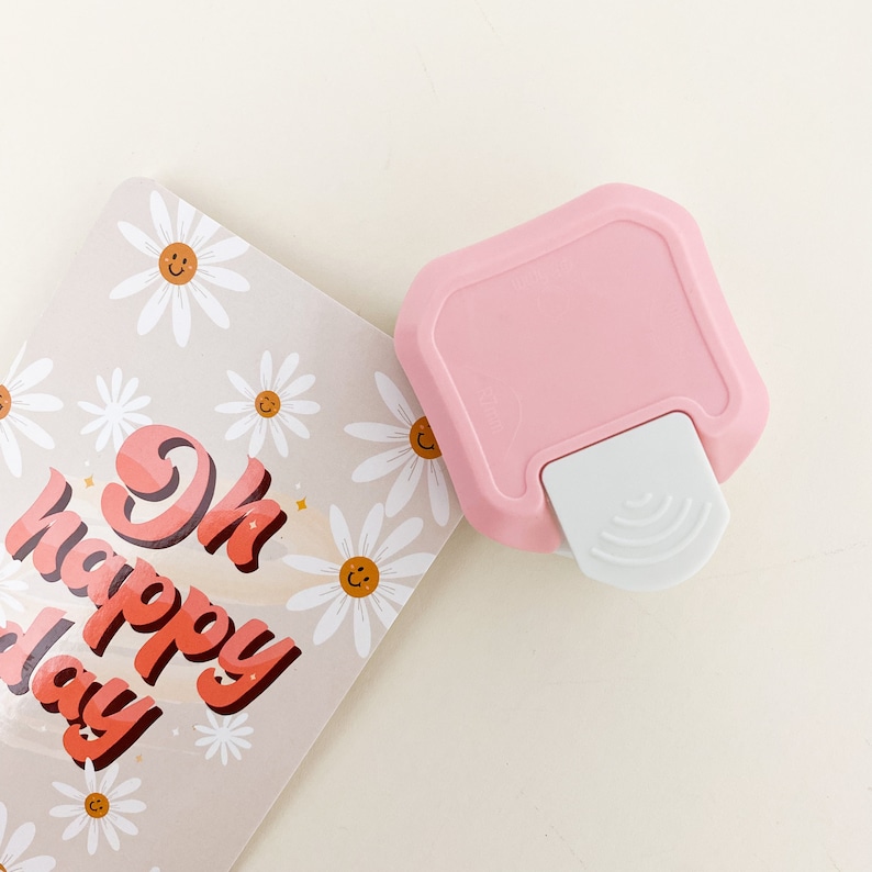 May include: A pink and white paper punch with a daisy design. The punch is on top of a card with the text "Ok Happy Day" in a retro font.