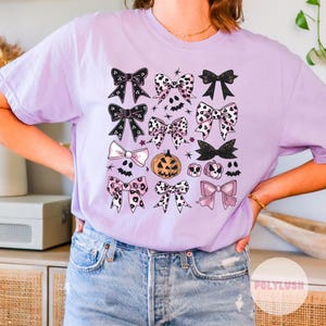 May include: A light purple t-shirt featuring a collection of bows in various designs, including polka dots and solid colors. Halloween-themed graphics like pumpkins and bats are also present. The shirt is a comfortable, casual style.
