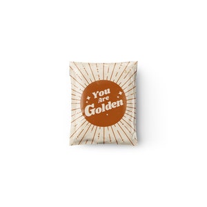Puede incluir: Un paquete color crema con un diseño de rayos de sol marrón y las palabras "You Are Golden" en una fuente retro. El paquete tiene una estética vintage.