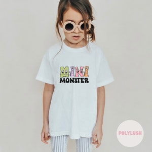 Könnte beinhalten: Weißes T-Shirt mit einem Aufdruck von bunten Monster-Cartoons, die "MINI MONSTER" buchstabieren.