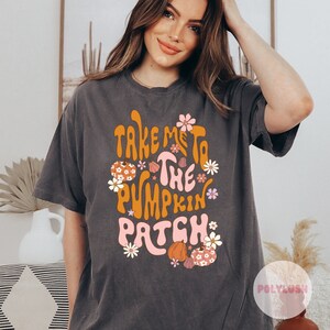Op de afbeelding: Een donkergrijs T-shirt met een retro-grafische afbeelding met de tekst "Take me to the pumpkin patch". De afbeelding is in oranje, roze en wit met bloemen en pompoenen.