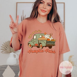 May include: A peach-coloured t-shirt featuring a vintage-style illustration of a green pickup truck filled with pumpkins. The text "Take me to the Pumpkin Patch" is printed in a retro font.