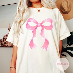Puede incluir: Una camiseta blanca con un diseño de lazo rosa en acuarela. El lazo es grande y está centrado en la camiseta.