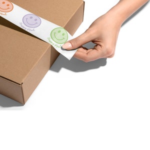 May include: A hand is peeling off a roll of white tape with a green smiley face and the text "Happy Mail" printed on it. The tape is being applied to a brown cardboard box.