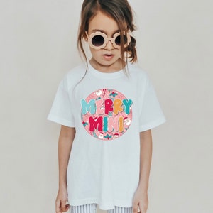 Könnte beinhalten: Ein weißes T-Shirt mit einem rosafarbenen Kreisdesign mit dem Text "Merry Mini" in bunten Blasenbuchstaben. Das Design enthält auch kleine Illustrationen von Weihnachtsmann, Zuckerstangen, Stechpalme und anderen Weihnachtselementen.