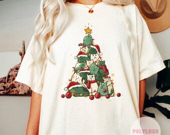 May include: A white t-shirt featuring a green Christmas tree design with a group of cats wearing Santa hats and scarves. The cats are stacked on top of each other, forming the shape of a tree. The tree is decorated with red and white baubles.