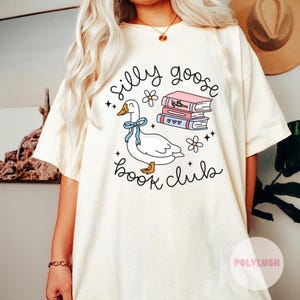 Pode incluir: Camiseta branca com um ganso de desenho animado segurando uma pilha de livros e o texto "silly goose book club".