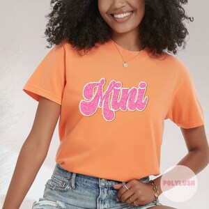 Könnte beinhalten: Ein leuchtend oranges T-Shirt mit einem pinkfarbenen und goldenen "Mini"-Motiv. Das Shirt ist aus weichem, bequemem Material und hat eine lockere Passform.