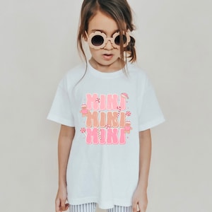 Könnte beinhalten: Weißes T-Shirt mit einer pinkfarbenen Grafik, auf der "Mini Mini Mini" mit Lebkuchenmännern und Zuckerstangen steht.