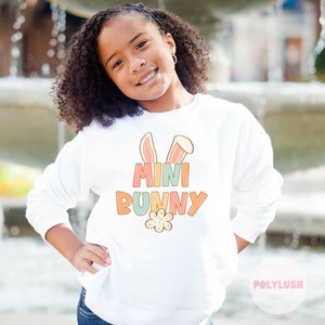 Könnte beinhalten: Weißer Pullover mit einer bunten Grafik von Hasenohren und dem Text "MINI BUNNY" mit einer Blume darunter.