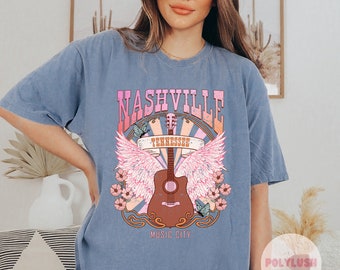 May include: A light blue t-shirt with a graphic print of a guitar with wings and the text "Nashville Tennessee Music City".