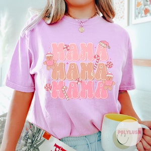Könnte beinhalten: Pinkes T-Shirt mit einem Grafikdesign des Wortes "Mama", das dreimal in einer Retro-Schrift wiederholt wird. Die Grafik enthält auch Lebkuchenmänner und Zuckerstangen.