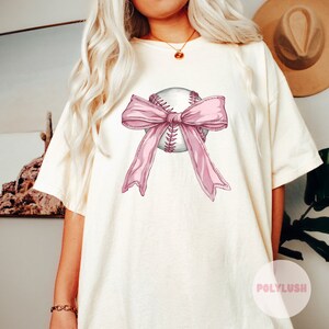 May include: Cream-coloured t-shirt featuring a baseball with a pink bow. The baseball is white with red stitching. The bow is a light pink colour. The shirt has short sleeves and a relaxed fit.