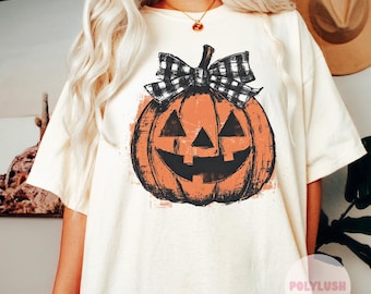 May include: A cream-coloured t-shirt with a distressed orange jack-o'-lantern graphic and a black and white plaid bow. The pumpkin has a smiling face. The shirt is a relaxed fit.