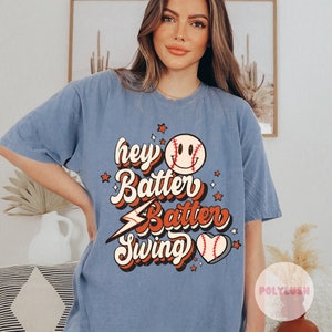 May include: A light blue t-shirt with a retro baseball graphic. The graphic features a smiling baseball with the text "hey batter batter swing".