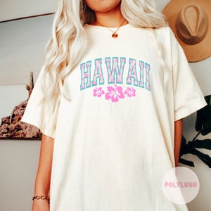 May include: Cream-colored t-shirt with the word "HAWAII" in a blue and pink floral design. Below the word are three pink hibiscus flowers. The shirt is a relaxed fit.