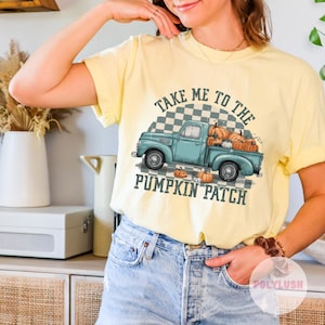 May include: Yellow t-shirt with a graphic of a teal pickup truck filled with pumpkins. The text "TAKE ME TO THE PUMPKIN PATCH" is arched above and below the truck. The shirt is a light yellow color.