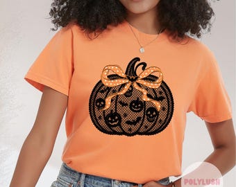 May include: Orange t-shirt with a black lace pumpkin design, featuring jack-o'-lantern faces and a bow with white stars. The shirt is short-sleeved and has a crew neck.