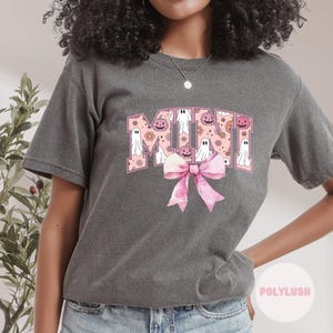 Könnte beinhalten: Dunkelgraues T-Shirt mit einem rosa-weißen Halloween-Design. Das Wort "MINI" ist mit Geistern, Kürbissen und floralen Akzenten verziert. Eine große rosa Schleife befindet sich darunter. Das T-Shirt hat einen lässigen Stil.