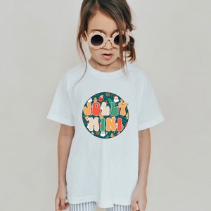 Könnte beinhalten: Weißes T-Shirt mit einer runden, bunten Grafik, auf der "Jolly Mini" steht, umgeben von weihnachtlichen Illustrationen.