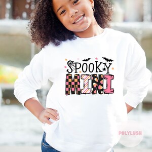 Könnte beinhalten: Weißer Pullover mit einem pinkfarbenen und schwarzen Aufdruck "Spooky Mini" mit einem Spinnennetz, Fledermäusen und Sternen.