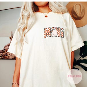 Könnte beinhalten: Cremefarbenes T-Shirt mit dem Wort "MINI" in einem karierten orange-weißen Muster. Ein Cartoon-Geist mit Blumenkrone befindet sich im "I". Das Shirt hat eine lockere Passform.