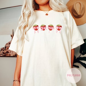Könnte beinhalten: Ein weißes T-Shirt mit einem Motiv aus fünf Erdbeeren, die mit rosa Schleifen gebunden sind. Der Text "POLYLUISH" ist in der rechten unteren Ecke des Bildes sichtbar.