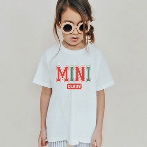 Könnte beinhalten: Weißes T-Shirt mit rotem, grünem und weißem Text, der "MINI CLAUS" lautet. Ein junges Kind trägt das Shirt und schaut in die Kamera.