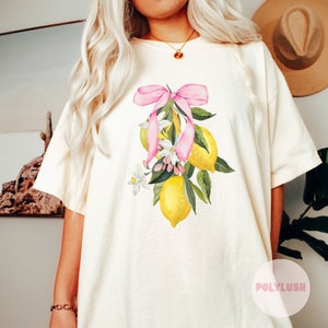 Pode incluir: Uma camiseta branca com um laço rosa e uma ilustração em aquarela de limões e flores. O texto "POLYLUISH" é visível no canto inferior direito da imagem.