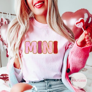 Könnte beinhalten: Pinkes T-Shirt mit dem Wort "MINI" in pinken, roten und goldenen Glitzerbuchstaben.