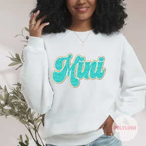 Könnte beinhalten: Weißer Pullover mit einem türkisfarbenen und goldenen Glitzer-Aufdruck "Mini".