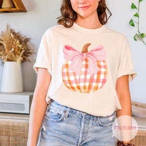 May include: A white t-shirt with a pink and orange plaid pumpkin with a pink bow on top. The pumpkin is centered on the shirt.