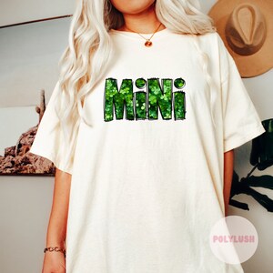 Könnte beinhalten: Cremefarbenes T-Shirt mit dem Wort "MINI" in Grün, gefüllt mit einem Kleeblattmuster. Der Text hat einen schwarzen Umriss. Das Shirt ist ein lässiger Stil, passend für St. Patrick's Day oder den Alltag.