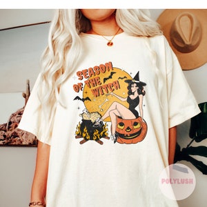 May include: Cream-coloured t-shirt with a retro Halloween graphic. The design features a witch sitting on a jack-o'-lantern, a cauldron, bats, and the text "Season of the Witch" in orange and black. The shirt has short sleeves.