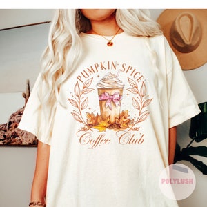 May include: Cream-colored t-shirt featuring a watercolor illustration of a pumpkin spice latte with whipped cream and a pink bow. The design includes fall leaves and the text "Pumpkin Spice Coffee Club."