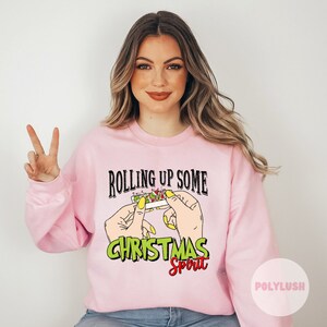 May include: Pink sweatshirt with the text "Rolling Up Some Christmas Spirit" and a graphic of hands rolling a joint with green and red marijuana.