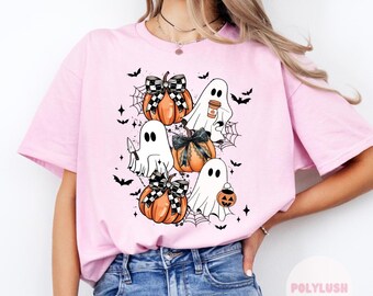 May include: Pink t-shirt featuring a graphic design of ghosts and pumpkins. The ghosts are holding a coffee cup, a knife, and a candy bucket. The pumpkins are decorated with black and white checked bows.