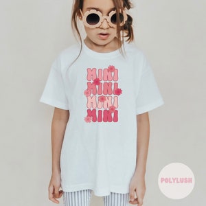 Könnte beinhalten: Weißes T-Shirt mit einer pinkfarbenen und roten Grafik, auf der viermal "mini" steht, mit pinkfarbenen Blumen um den Text herum.