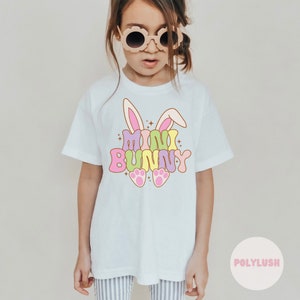 Könnte beinhalten: Weißes T-Shirt mit einer bunten Grafik eines Hasen mit dem Text "mini bunny". Der Hase hat rosa Ohren, einen hellgrünen Körper und rosa Pfotenabdrücke.