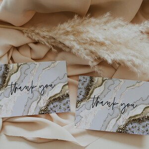 May include: Two folded cards with a marble design and the words "thank you" in black script. The cards have a light blue and gold color scheme. The background is a beige fabric with dried ornamental grass.
