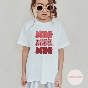 Könnte beinhalten: Weißes T-Shirt mit dem Text "MINI MINI MINI" in Rosa und Rot mit Herzen. Der Text ist in einer Retro-Schriftart.